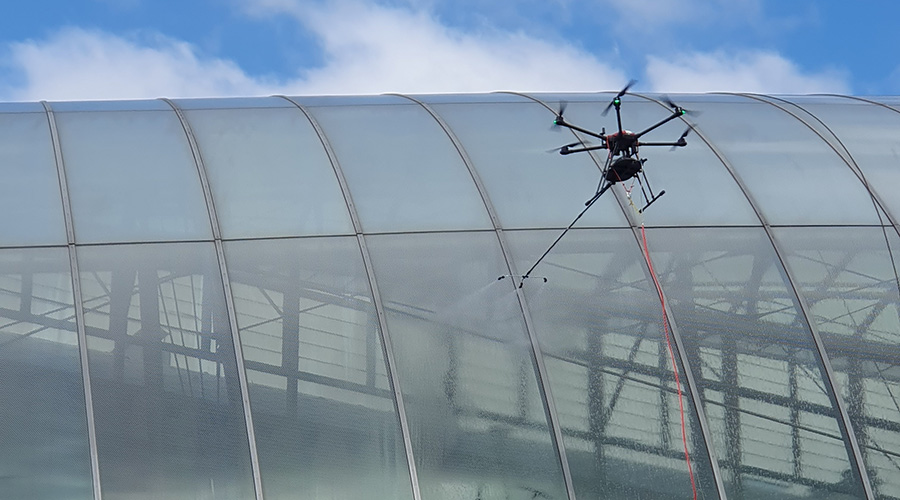 Een drone die met spuitnevel een groot gebogen glazen dak reinigt, gefotografeerd vanaf de grond