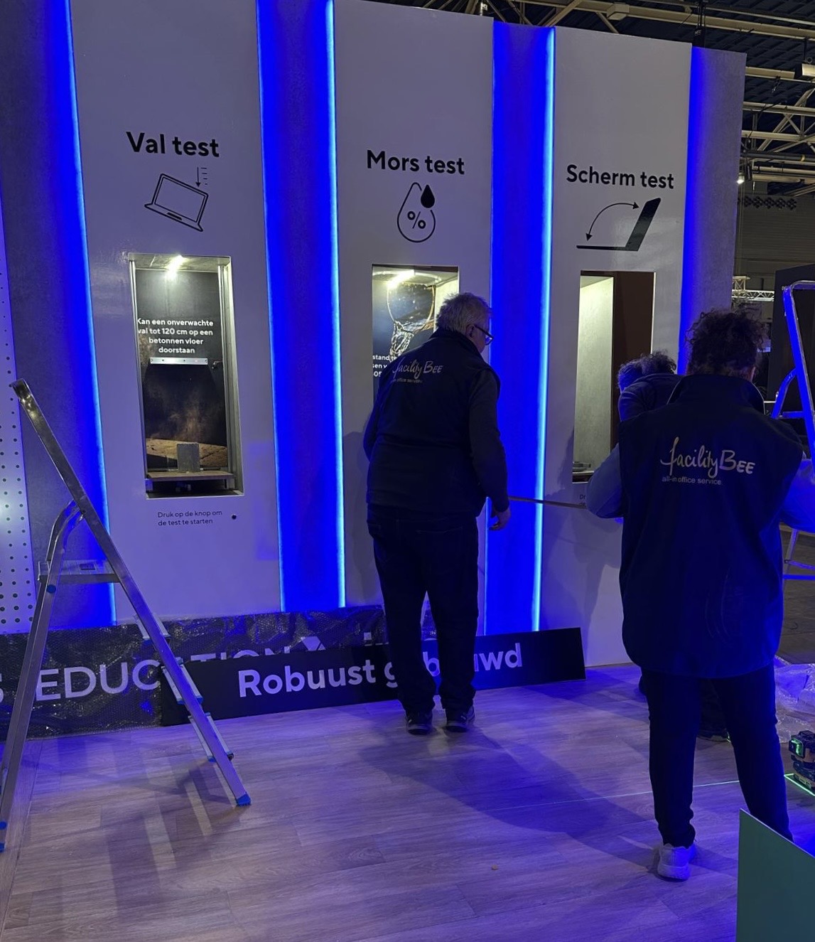 Twee FacilityBee-medewerkers in blauwe vesten bouwen een witte beurswand met drie teststations op in een evenementshal