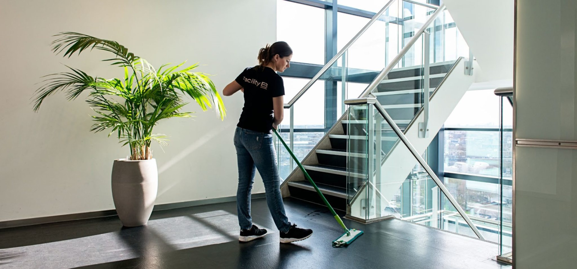 Een FacilityBee-medewerkster die met een groene mop een donkere vloer op de bovenverdieping van een kantoor poetst, met een trap en glazen gevel achter haar